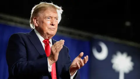 US President Donald Trump standing on a stage dressed in a navy suit and red tie clapping to an audience not in view. 
