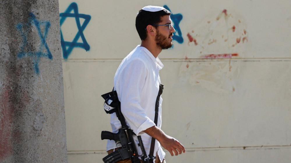 An Israeli settler in a white shirt and carrying a gun walks in front of a wall