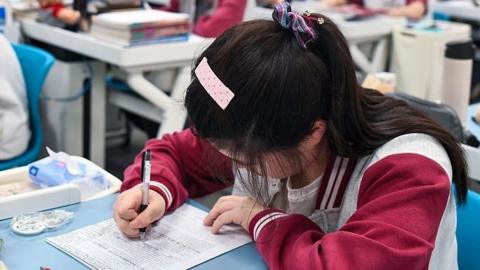 A teenager sits at a desk with her head down looking at a paper. She is writing on it with black pen. Stationery and a teddy bear sits on her desk. In the background are other teenagers at desks, all spaced out, in a classroom.