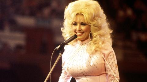 Dolly Parton smiles on stage wearing a white rhinestone jumpsuit.
