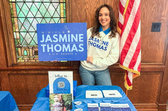 N’Kiyla Jasmine Thomas stands in front of a campaign table while holding a blue sign that reads "N’Kiyla Jasmine Thomas, U.S. Senate"