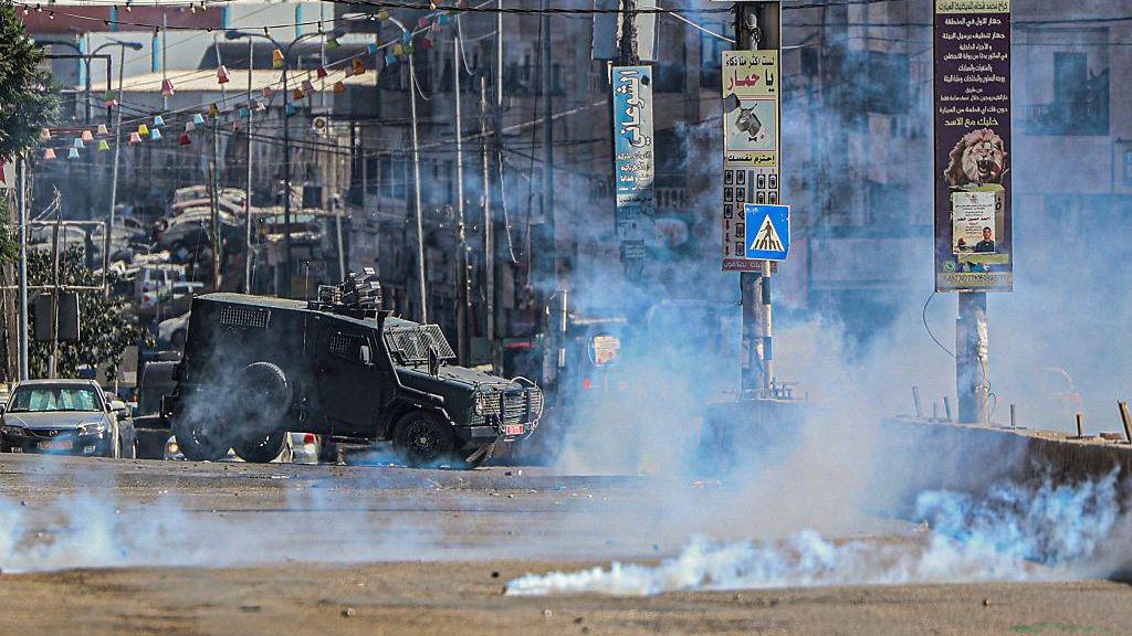 An Israeli armoured vehicle moves along a street as tear gas fills the air, during a raid in Qalandia refugee camp, in the occupied West Bank (25 August 2026)