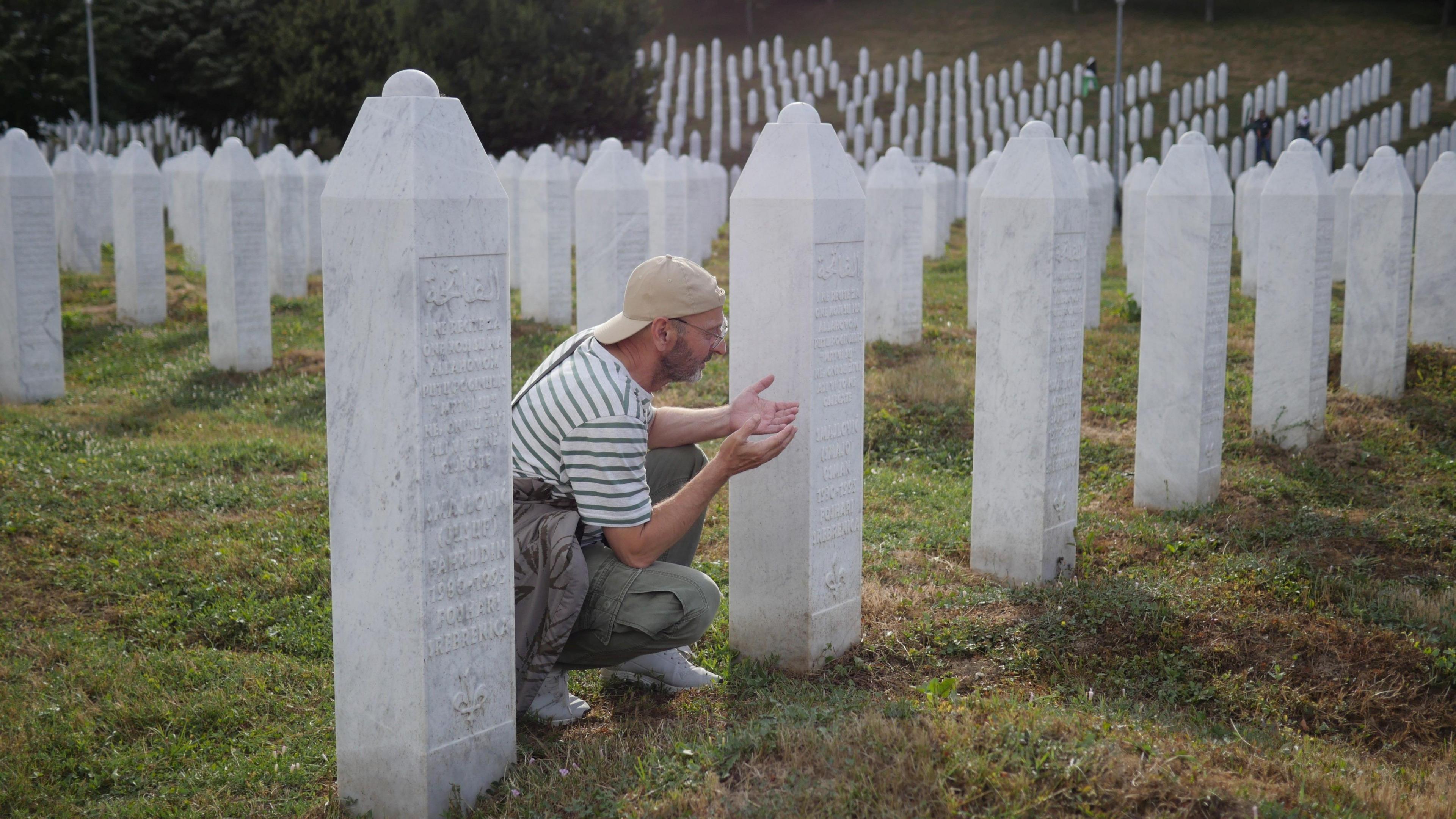 A recent photograph of a man in a modern outfit (striped t-shirt, cargo trousers and a backwards cap) prays with his palms facing upwards next to a plain white grave. The cemetery is filled with thousands of identical graves where victims of the Srebrenica massacre are buried.