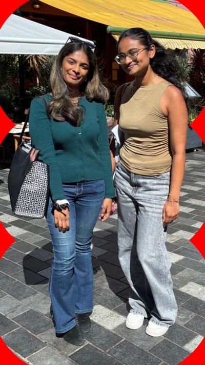 Two women with long dark hair stand in an outdoor square. One wears a turquoise top, the other wears a sleeveless khaki top
