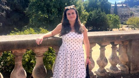 Ella Kipling, a young woman with long dark hair, smiles at the camera. She is wearing a long, white dress with black polkadots and is standing in front of a large park with old buildings in the background.