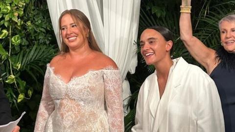 Two women hold hands in white bridalwear; one wears a white tailored suit, while the other wears a lace dress.