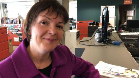 Elizabeth in a purple jacket sits at a desk in an open-plan office. Red filing cabinets, shelves of books, computer monitors and office equipment are visible in the background, with papers and magazines on the desk in the foreground.