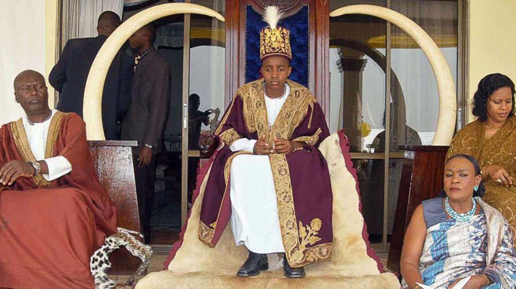 King Oyo at 13 sitting on a throne in a red robe and embroidered crown. Two elephant tusks can be seen wither side