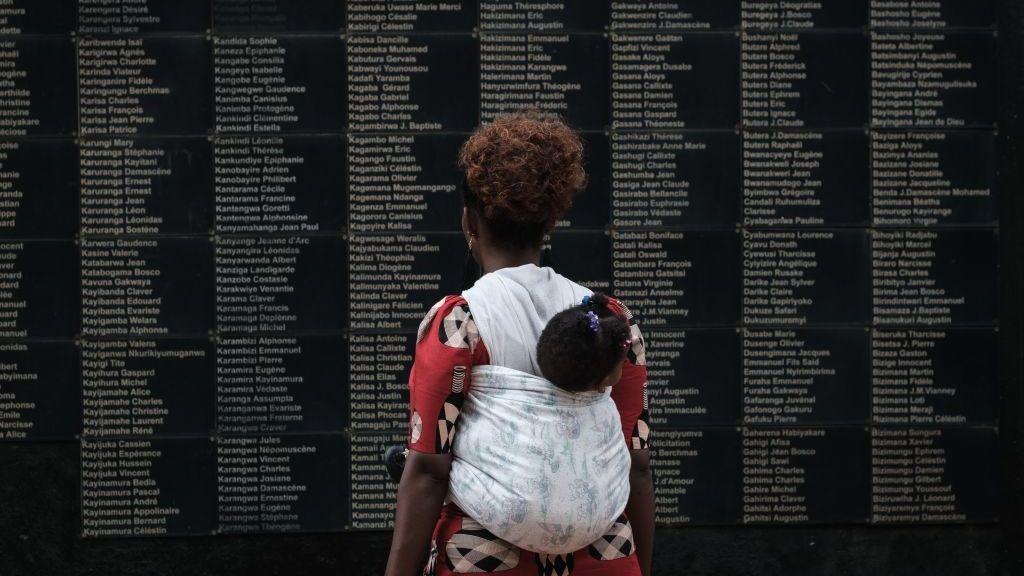 A woman with a baby on her back faces away from the camera. She is looking at a wall with names etched into it.