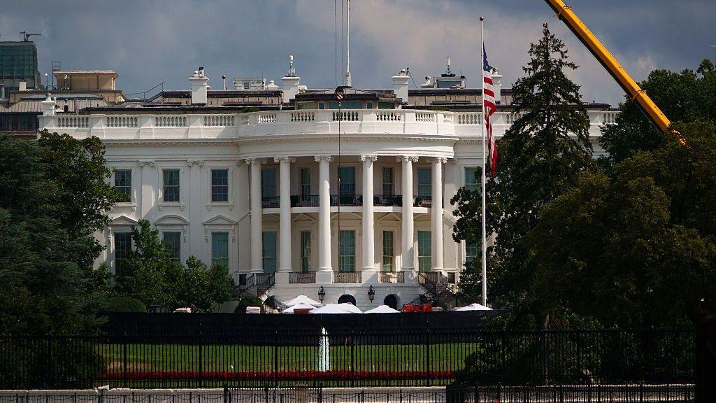 Exterior shot of the White House's South lawn, the Truman balcony (portico area enclosed by six white columns in the Ionic style, with a distinctive scroll-shaped ornaments called volutes on the capital. A yellow crane can be seen emerging from trees to the left of the frame