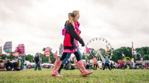 Two people walk through a festival site wearing waterproofs and wellies