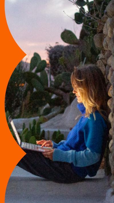 A woman with shoulder-length blonde hair sits outside with a laptop on her knee