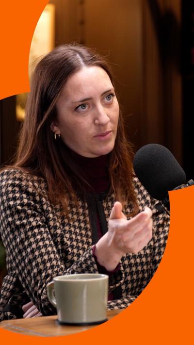 A woman with brown hair sits in front of a microphone