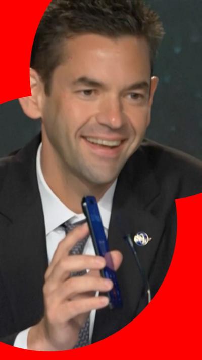 A man smiles as he holds up a phone through which the US president is addressing the audience