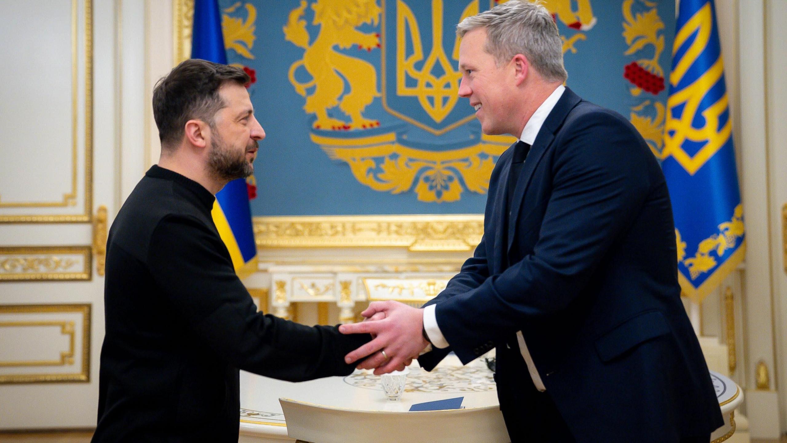 Volodymyr Zelensky, in black mockneck, puts hand forward, which Dan Driscoll, which grey hair and navy suit, grips in both hands. In the background are gold and blue insignia and flags