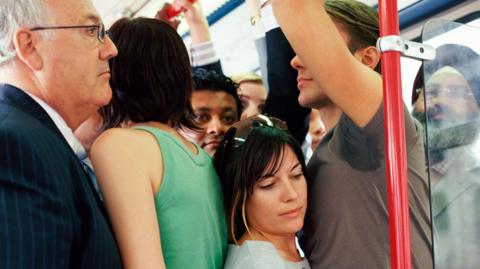 A shot of a crowded train. A man in a pinstriped suit stands next to a woman wearing a green top. A smaller woman is standing next to a red pole, next to her a man in a brown t-shirt, with sweat patches visible at his armpit. 