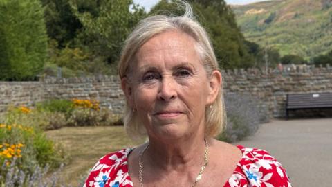 Janett wears a red floral dress and gold necklace. She has short light blonde hair and is smiling. It is a head and shoulders shot of her.