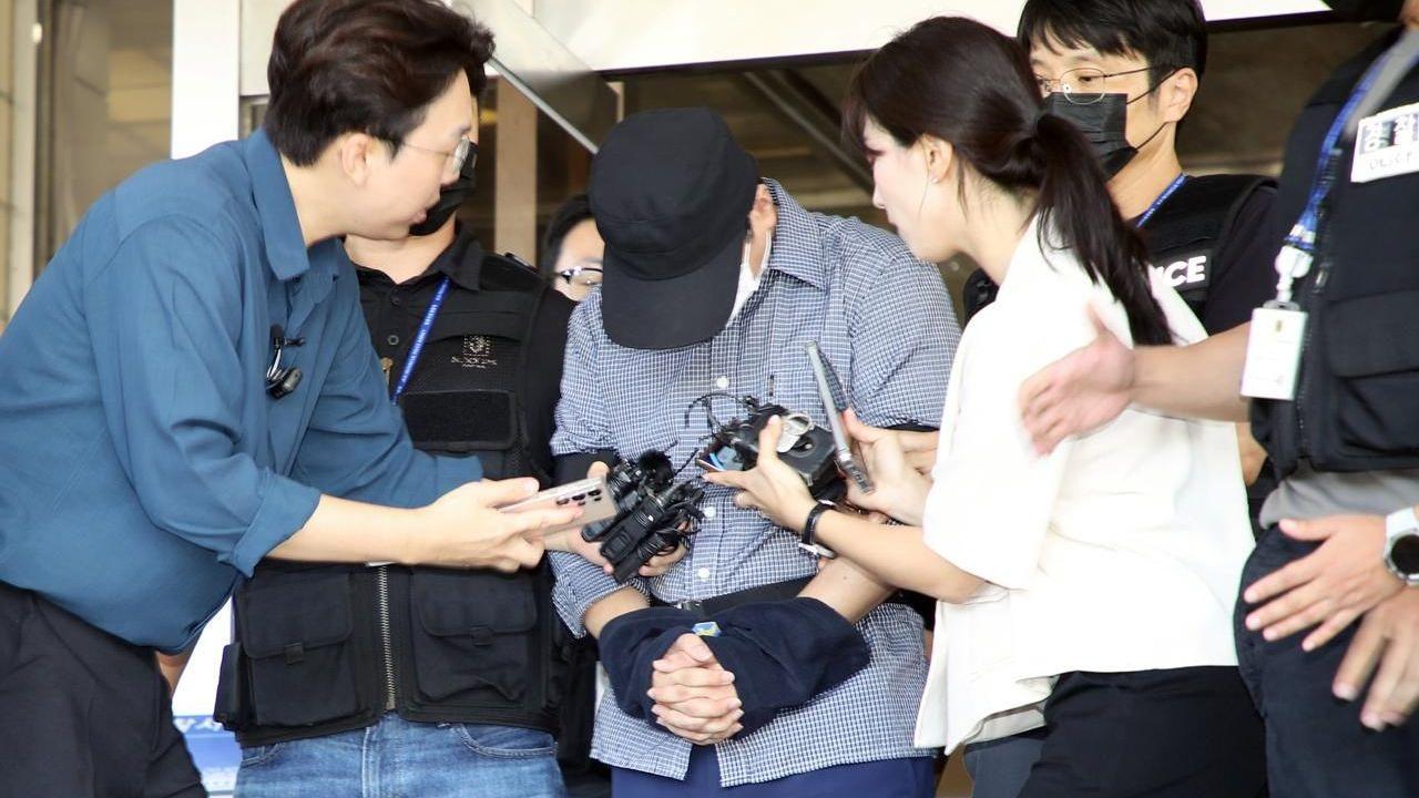 A man wearing a navy blue cap and a face mask bows his head as he is questioned by reporters