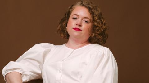 Woman with curly red hair and bright red lipstick faces the camera. She is wearing a white shirt