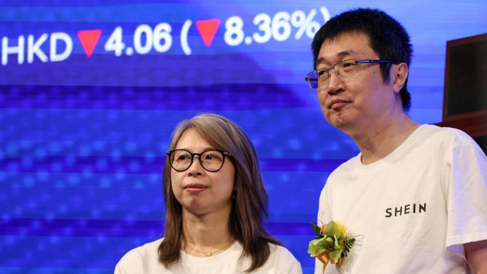 Shein's founder, Xu Yangtian (right), stands on a stage next to financial director Poppy Bao during the company's listing ceremony. Behind them is a blue screen showing Shein's shares down by 8.36%. 