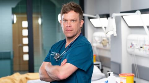 Promotional image of William Beck in character as Dylan Keogh. He is photographed from the waist up and is wearing blue medical scrubs inside the set of the emergency department. A stethoscope can be seen around his neck. Beck is stood to the side with his arms crossed. He is looking at the camera and is not smiling. 