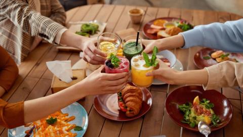 Close up of four women putting their drinks together in a 'cheers' motion. They are sat around a wooden table with plates of food in front of them.