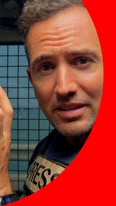 A man with short grey hair looks at the camera. He is wearing a blue flak jacket.