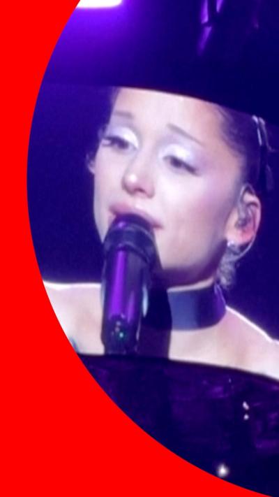 A woman is projected on a screen speaking into a microphone, with purple lights behind it.
