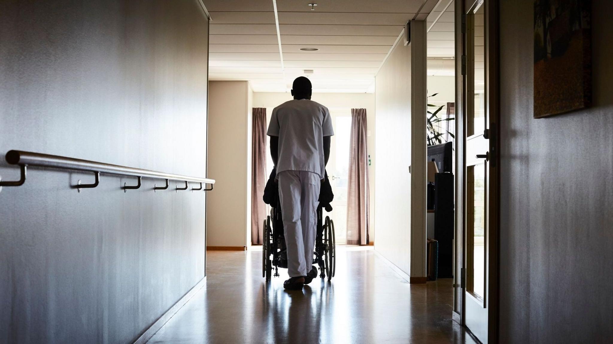 Man pushing resident of a care home down a dimly-lit hallway, their backs towards the camera