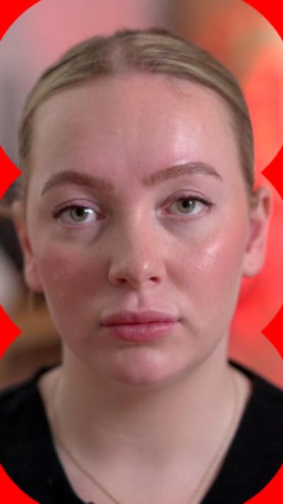Ffion looks at the camera with a stern expression. She wears a black uniform. She has her blonde hair in a slick back bun and wears a gold necklace. She is sat in front of a work place which has a sink on, alongside plants and a red mood light.