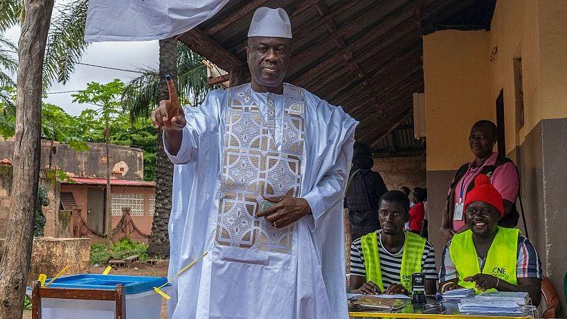 Gen Horta N'Tam in a white traditional outfit holds up his ink-stained finger after voting at an outdoor polling station in Bissau, as election officials sit beside ballot materials