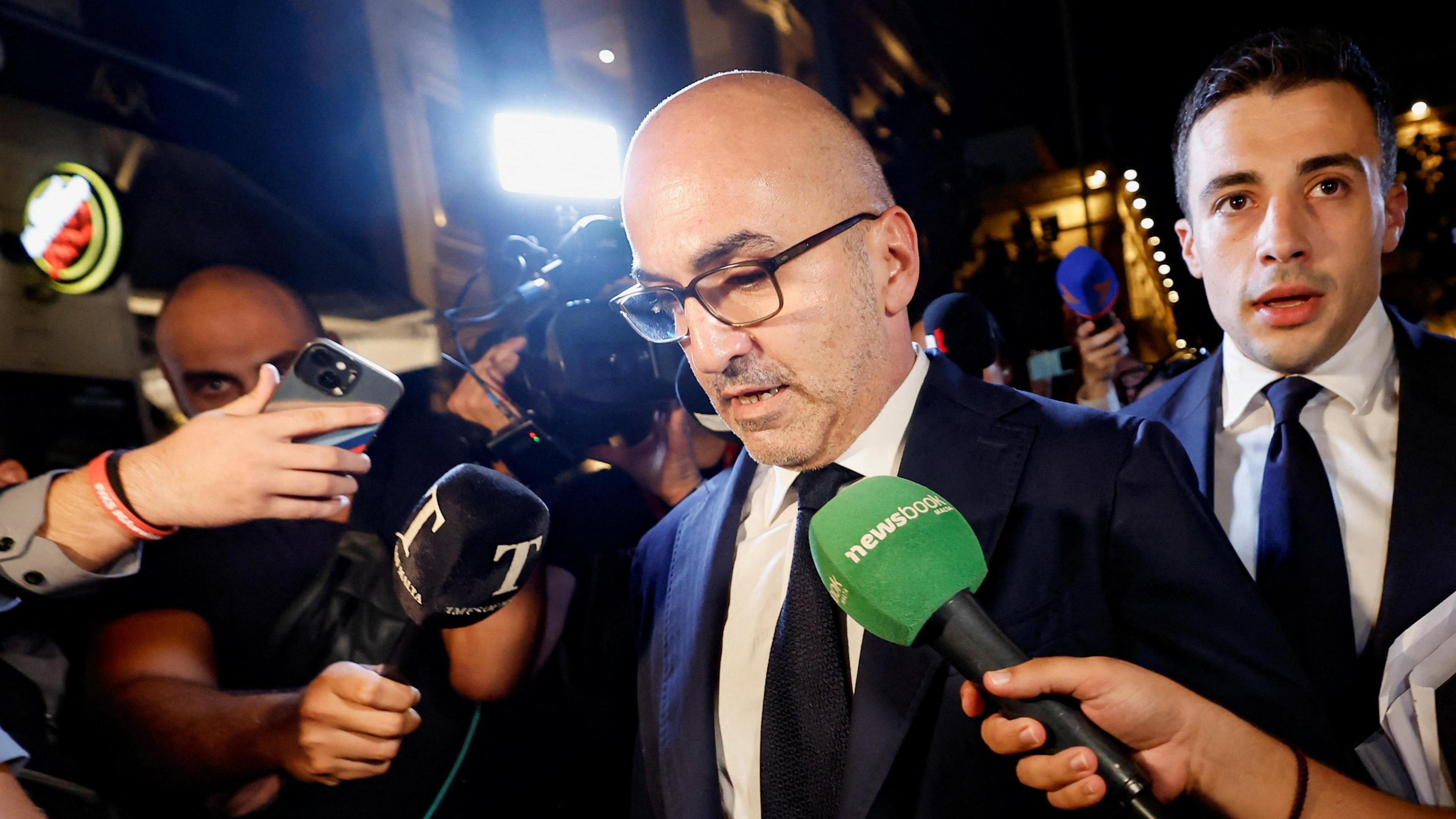 Yorgen Fenech wears a black suit and glasses surrounded by journalists