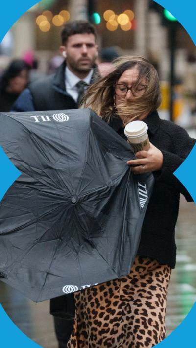 A woman in a leopard print dress and navy jacket struggles in windy weather with an umbrella and a portable coffee cup. A man behind her is wearing a navy puffer jacket.
