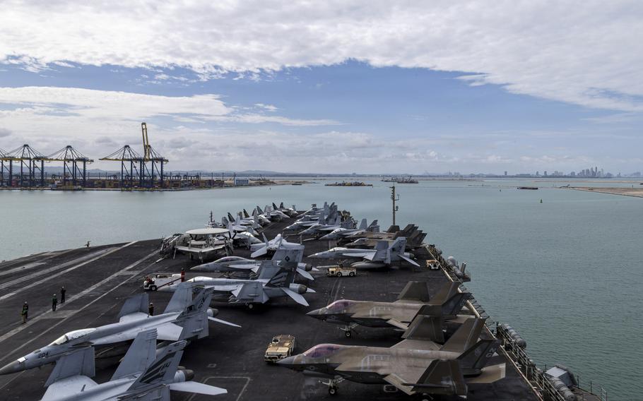 Fighter jets sit parked on the deck of an aircraft carrier in the foreground, with the water of the port extending towards the horizon line in the distance.