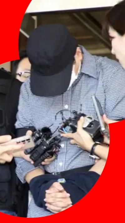A man in handcuffs hands his head. He is wearing a navy blue baseball cap and a white face mask. His shirt has a small-grained pattern in blue and white.