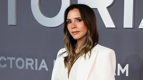 Victoria Beckham wearing a white top and standing in front of a dark grey panel showing her name