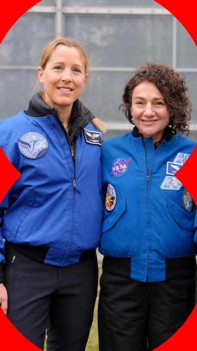 Two women, one with straight ginger hair, and the other with curly brown hair smile at the camera. They are both wearing navy trousers and bright blue zip-up jackets with space flight insignia on.