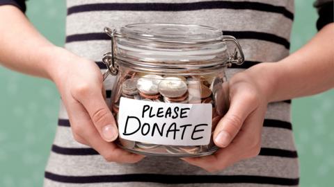 A shot of someone's torso, in a black and white striped top, holding a glass jar with coins in it. On the front is a label that reads: Please donate. 
