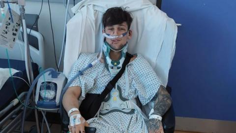 Liam McCracken, who is wearing a patterned hospital gown, is sitting in a chair next to a hospital bed. He is wearing a neck brace and is surrounded by medical equipment. 