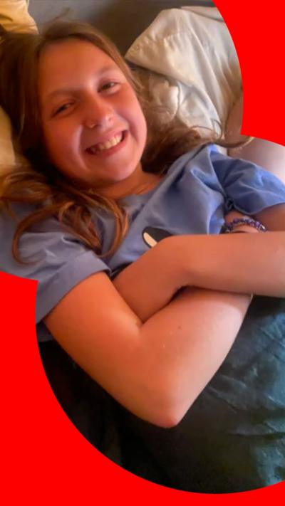 A teenaged girl with long hair is lying on a couch with her arms folded. She is smiling, wearing a blue T-shirt with a logo on it and is lying under a blanket.