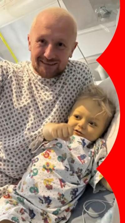 A man and baby smile at the camera, they wear hospital gowns