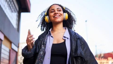 A young Black woman smiles as she walks down the street listening to music on her phone, while wearing large yellow headphones
