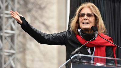 Gloria Steinem, in a red scarf and sunglasses, gestures with an outstretched arm as she speaks at a podium