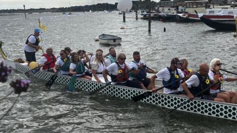 Liz Masding and Mike Markham-Lee are sitting on the long black and white dragon boat. There are multiple rowers holding oars on the boat. 