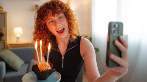 A women hold a small birthday cupcake with candles and takes a selfie 