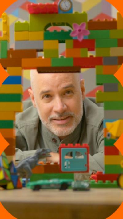A man with a short, grey beard looks through a bridge made of different coloured plastic toy bricks