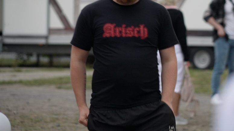The torso of a man wearing a black t-shirt which says 'Arisch', German for Aryan written in a red Gothic-style font.