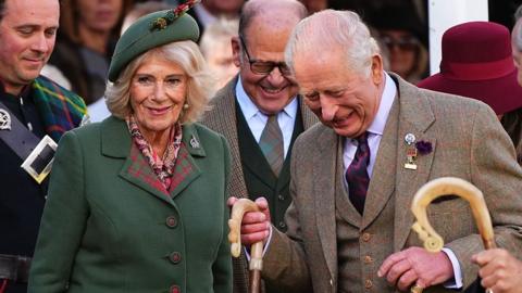 Queen Camilla, wearing a green blazer and a green hat, next to King Charles who is wearing a tweed three-piece suit. They are surrounded by officials and are in high spirits at the Braemar Gathering
