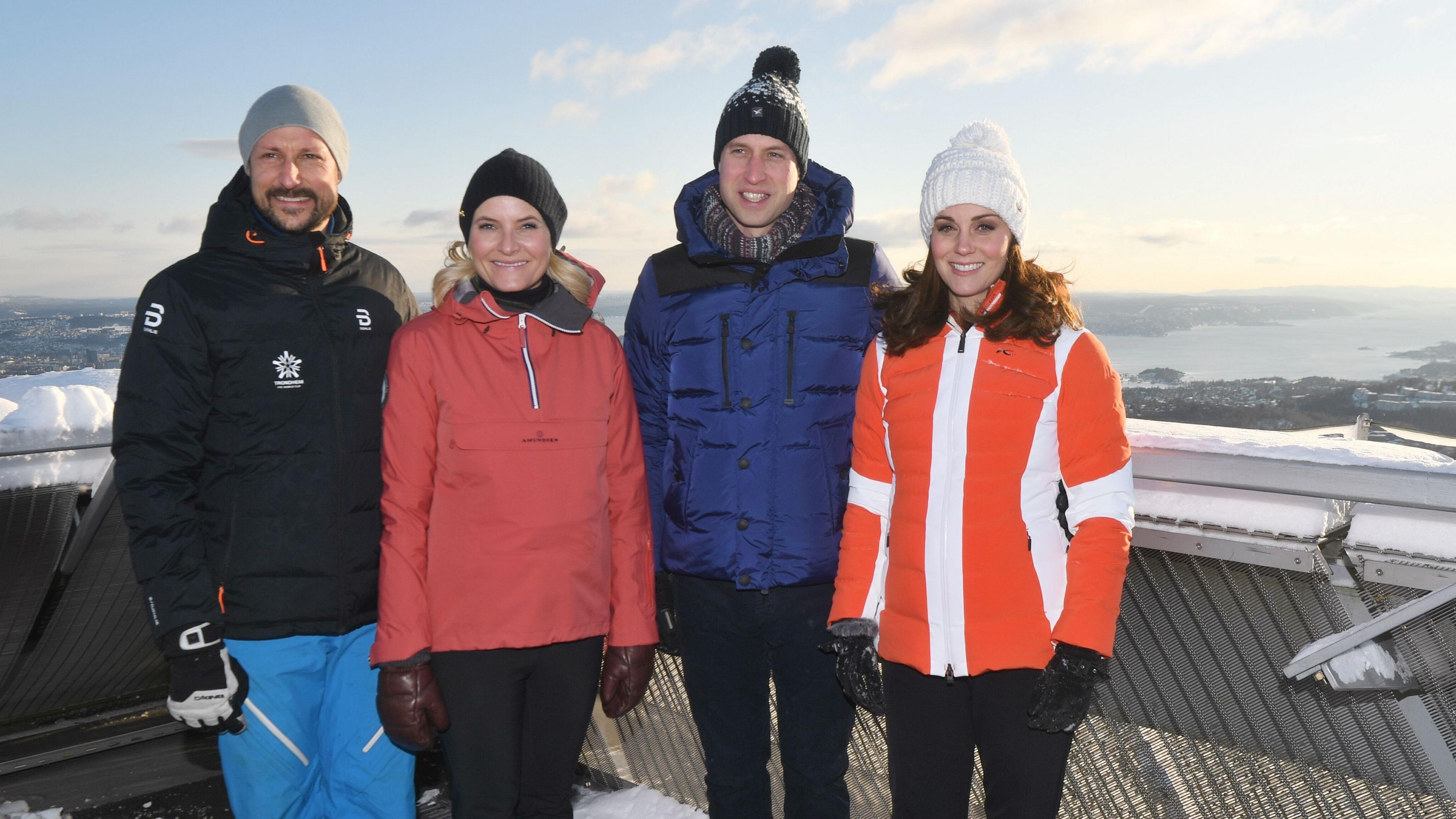 Four people stand smiling at the camera in ski jackets and woollen hats. On the left is a man with a goattee beard and next to him is a woman with blonde hair. Next is Prince William wearing a blue ski jacket and pom-pom hat, and Kate wearing a bright orange jacket and white hat. They are standing outside and look to be high-up on a mountain, and in the background is snow and a view.