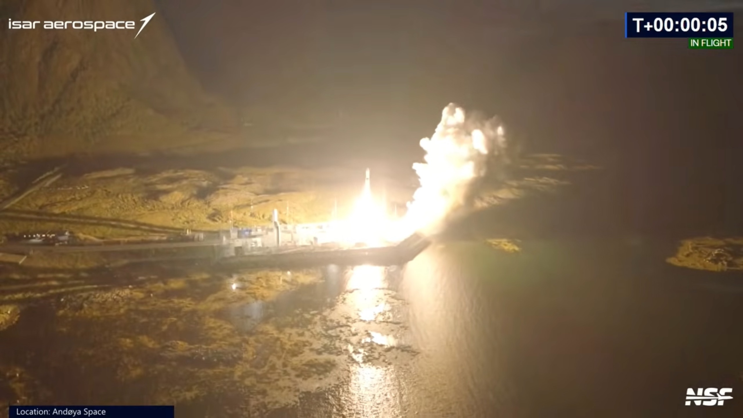 a white rocket launches into a dark night sky from a seaside pad with mountains in the background